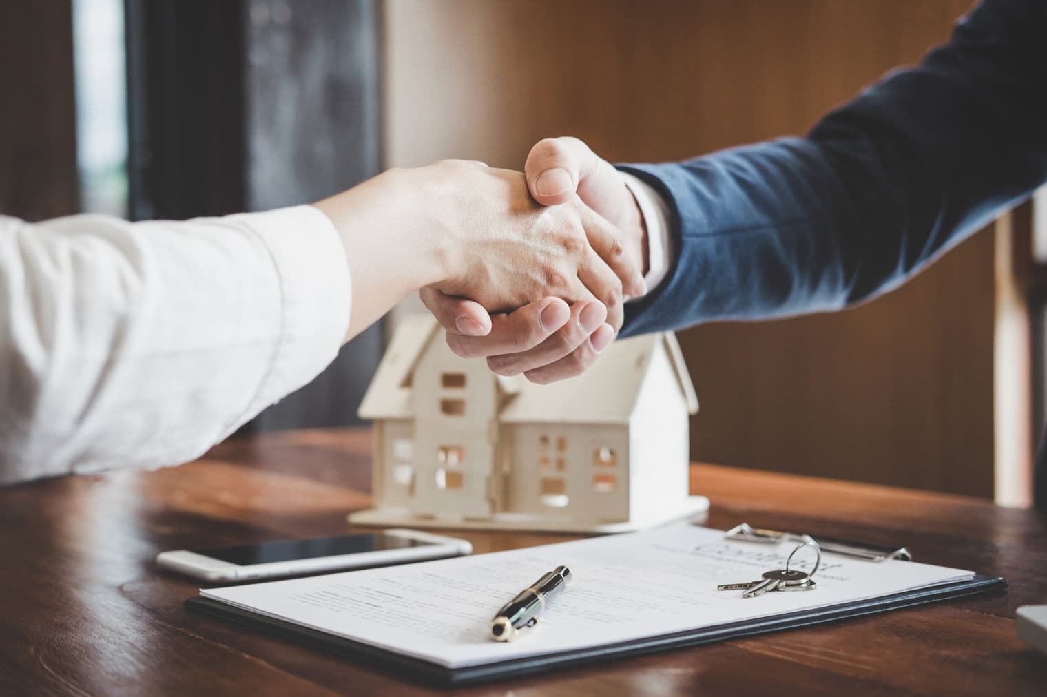 Close-up of a handshake over a wooden table with a house model, keys, pen, phone, and contract.