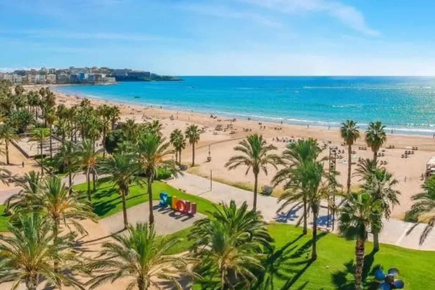 Vista frente a la playa de Salou, España, con palmeras, playa de arena, mar azul y horizonte de la ciudad bajo un cielo azul despejado.