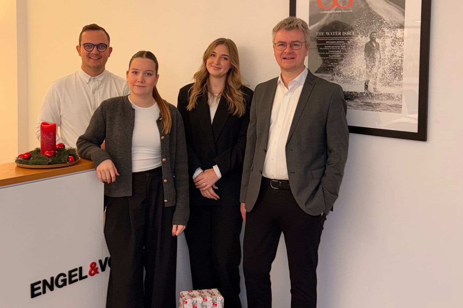 Vier Engel & Völkers-Makler posieren vor einem weißen Tresen mit Geschenken. An der Wand hinter ihnen hängt ein gerahmtes Zeitschriftencover.