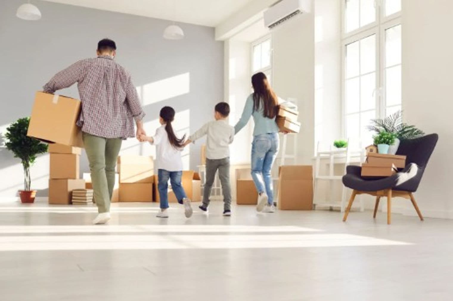 Familie zieht in ein neues Zuhause. Eltern tragen Kisten, Kinder halten Händchen und betreten einen hellen, leeren Raum voller Umzugskartons.