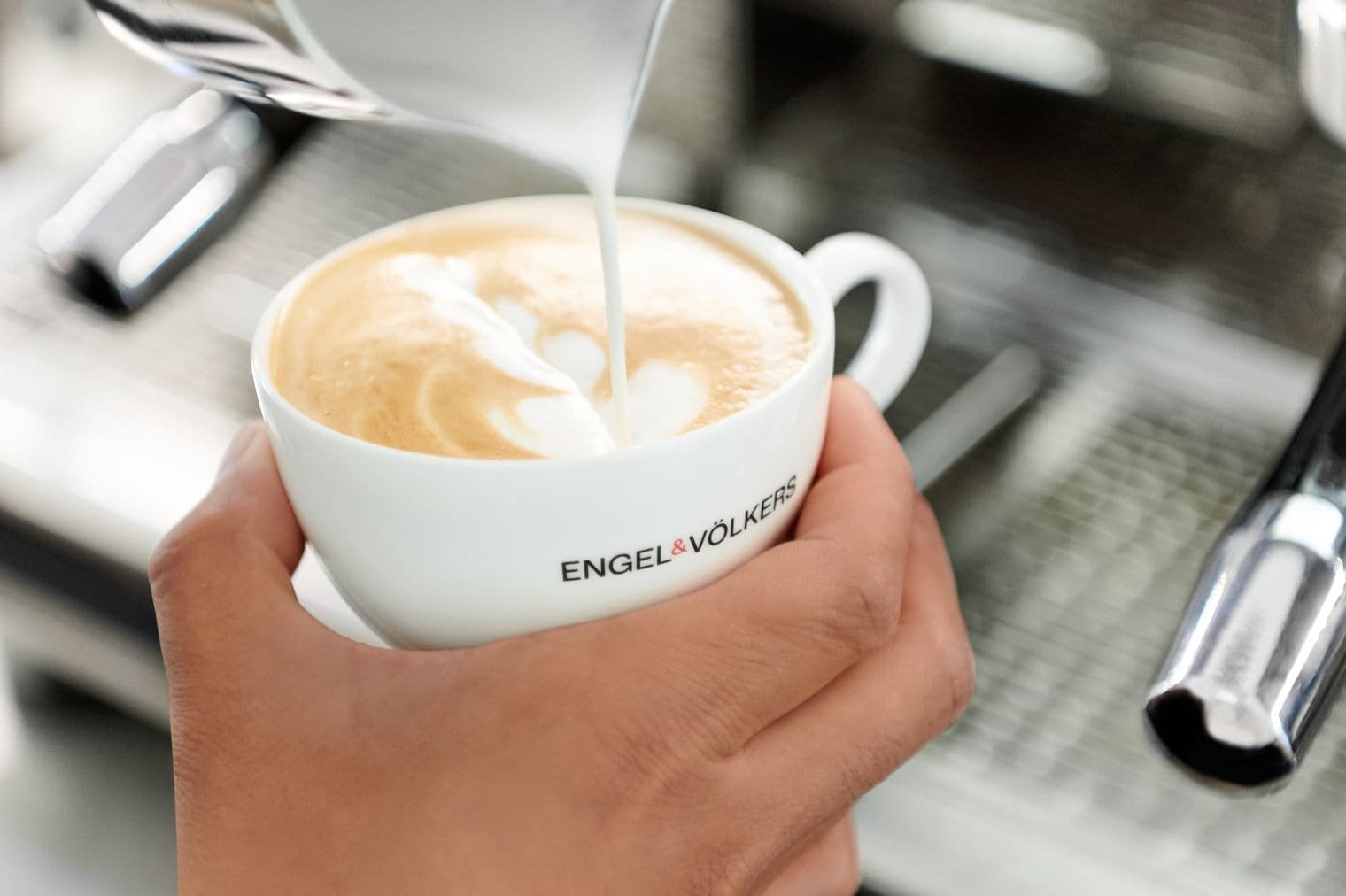 Nahaufnahme von Milch, die in eine Engel & Völkers Kaffeetasse gegossen wird und Latte Art erzeugt.