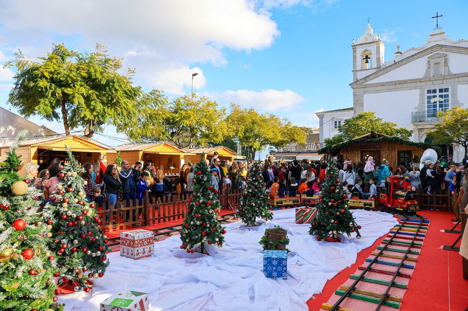 Mercado de Natal ao ar livre com árvores decoradas, caixas de presentes e um trem de brinquedo sobre um tapete vermelho. Pessoas observam barracas de madeira sob um céu azul.