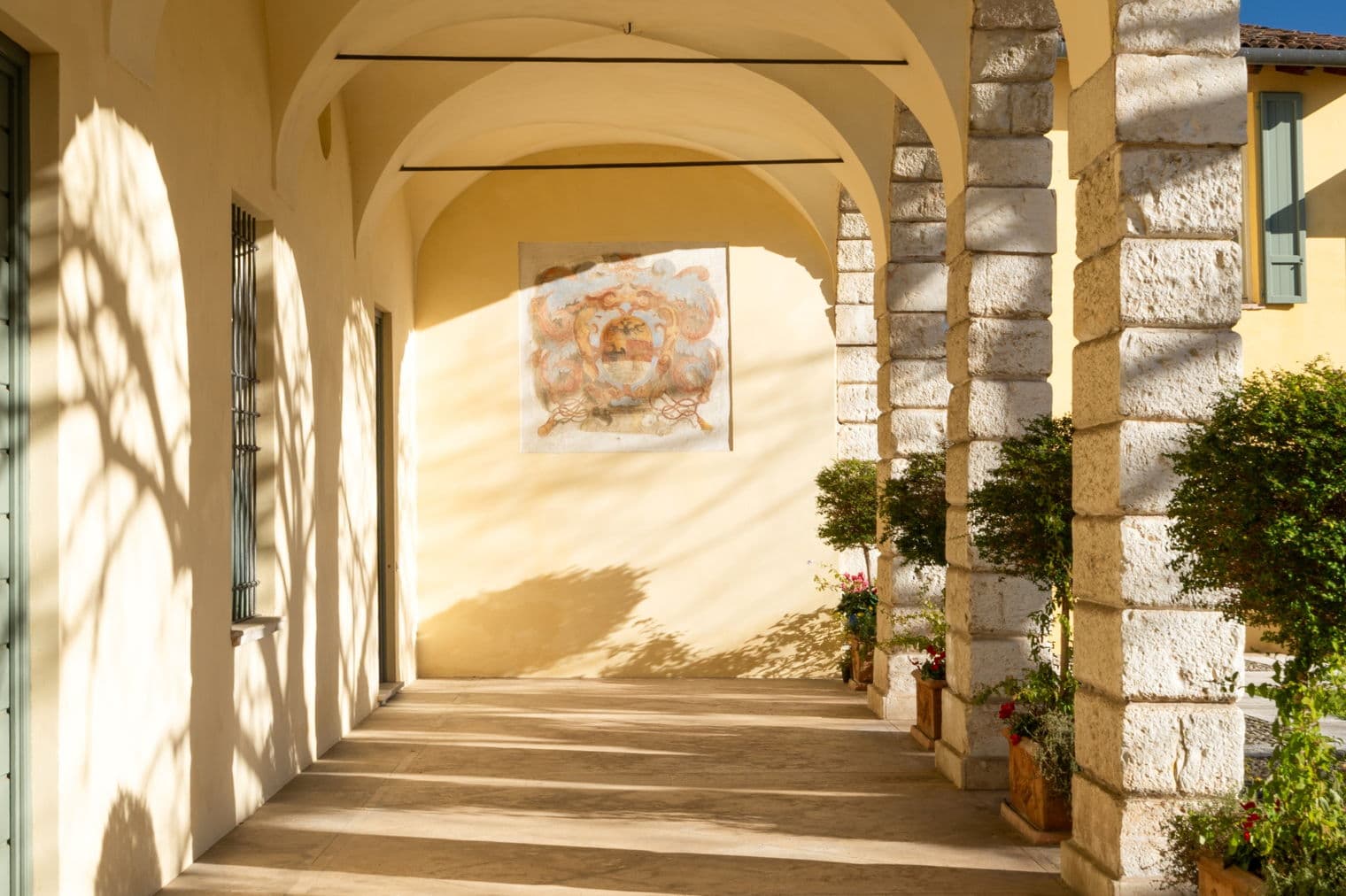Überdachter Gehweg mit Steinsäulen, gewölbten Decken und Topfpflanzen. Ein gemaltes Wappen schmückt die gelbe Wand. Sonnenlicht wirft Schatten auf den Steinboden.