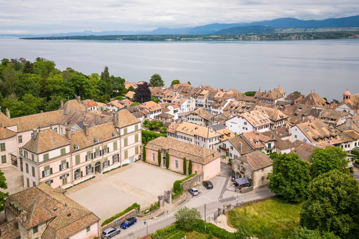 Vista aerea di una città europea con un grande lago e montagne sullo sfondo. La città ha molti edifici con tetti marroni e un grande castello rosa.