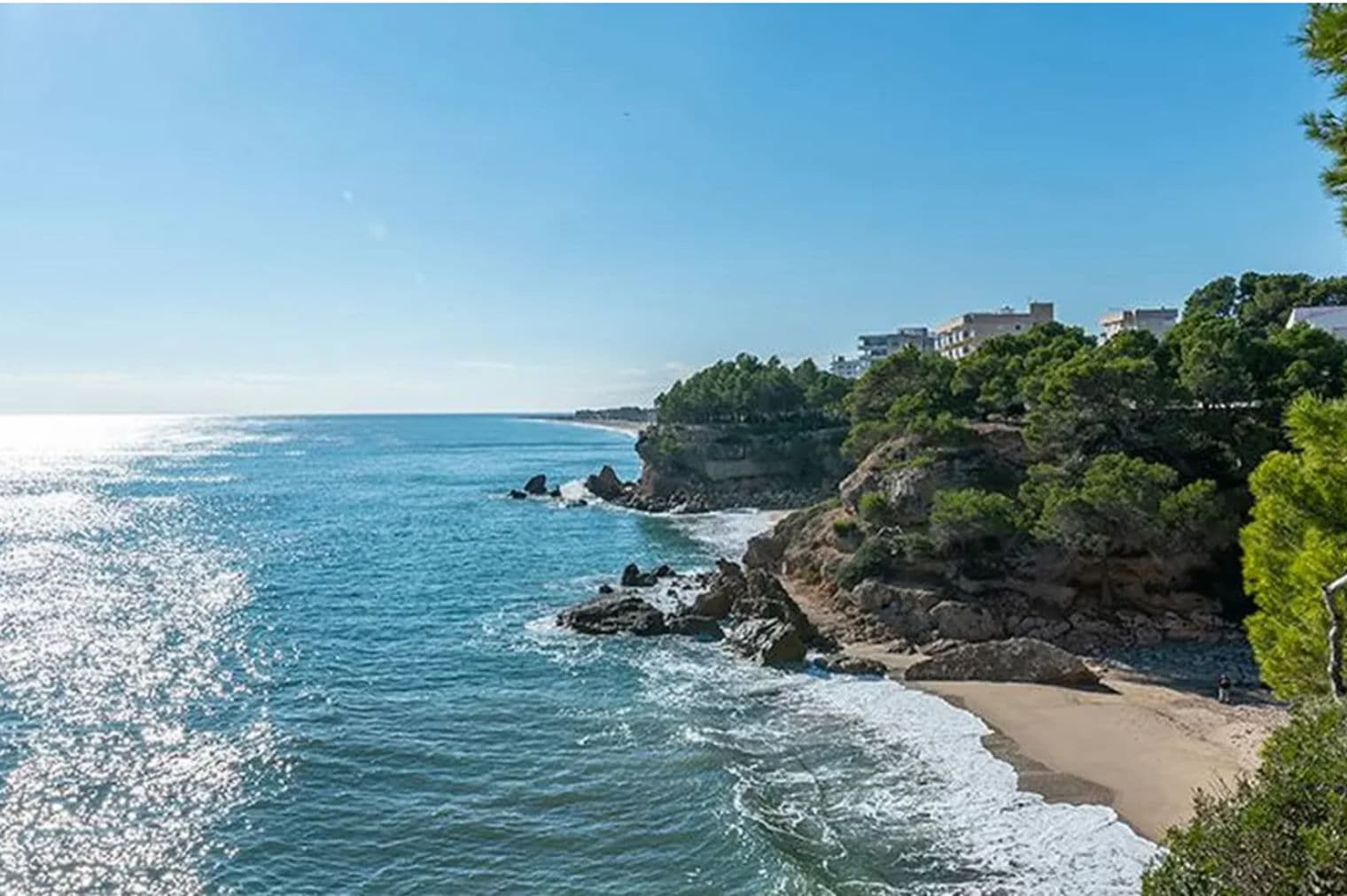 Vista al océano con acantilados rocosos, árboles y edificios bajo un cielo azul despejado. Olas rompen en una playa de arena.