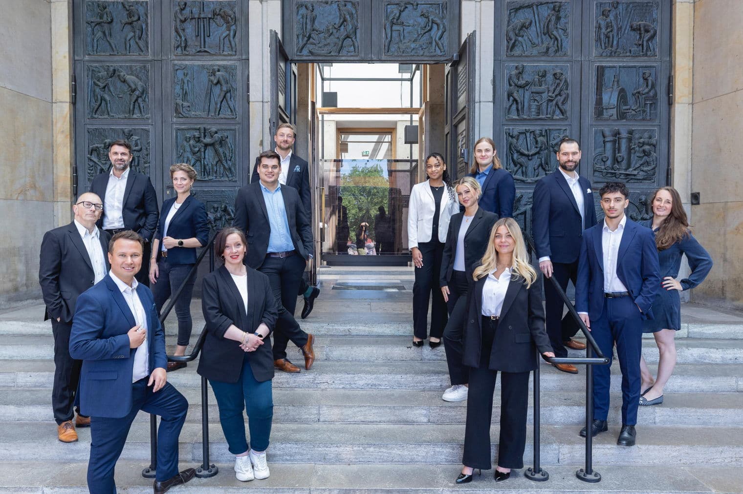 A group of real estate agents in business attire pose on stone steps in front of a large, ornate bronze door.