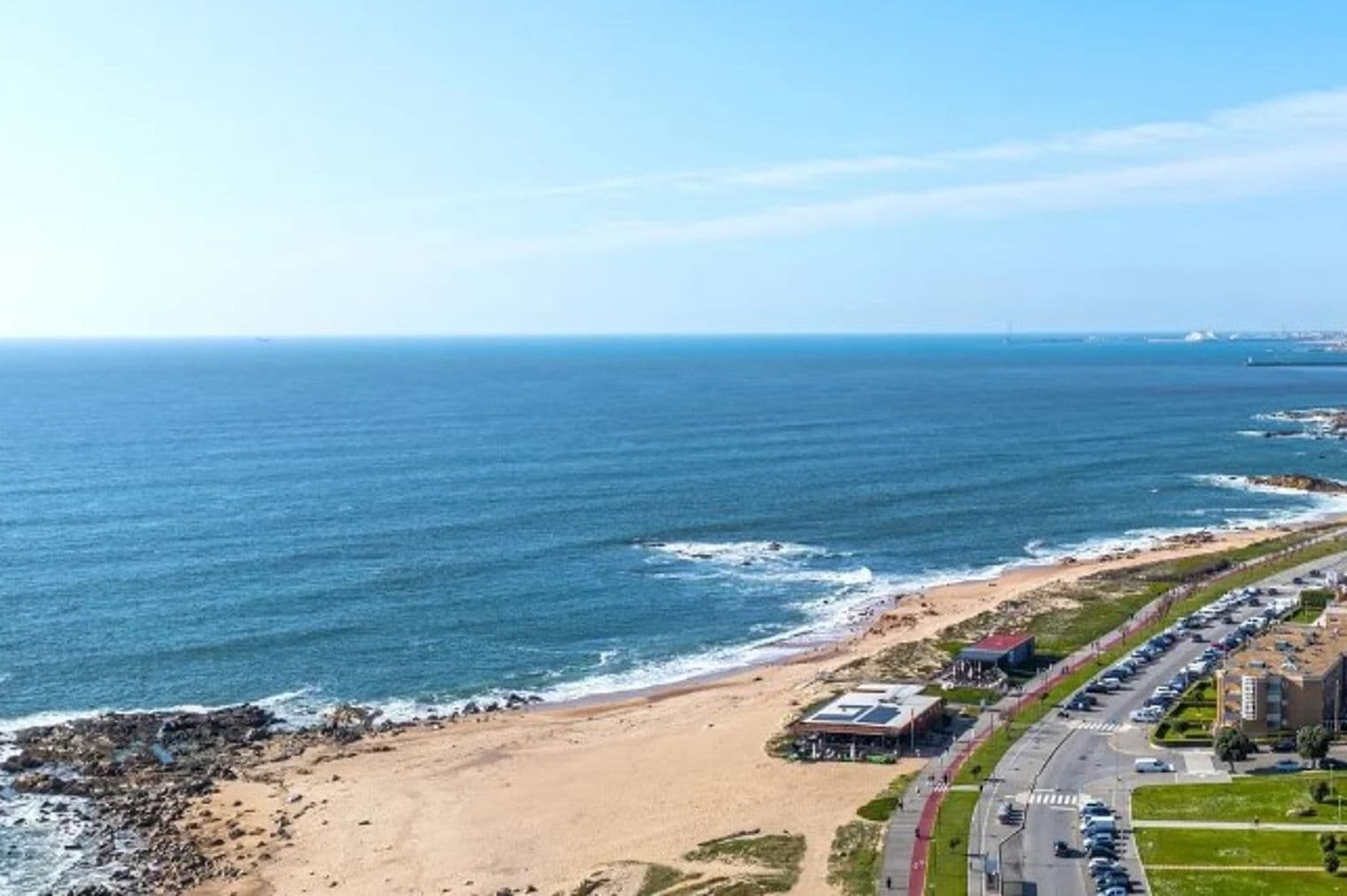 Meerblick auf Porto, Portugal. Sandstrand trifft auf blaues Wasser, mit Stadtgebäuden und einer Straße mit Autos auf der rechten Seite.