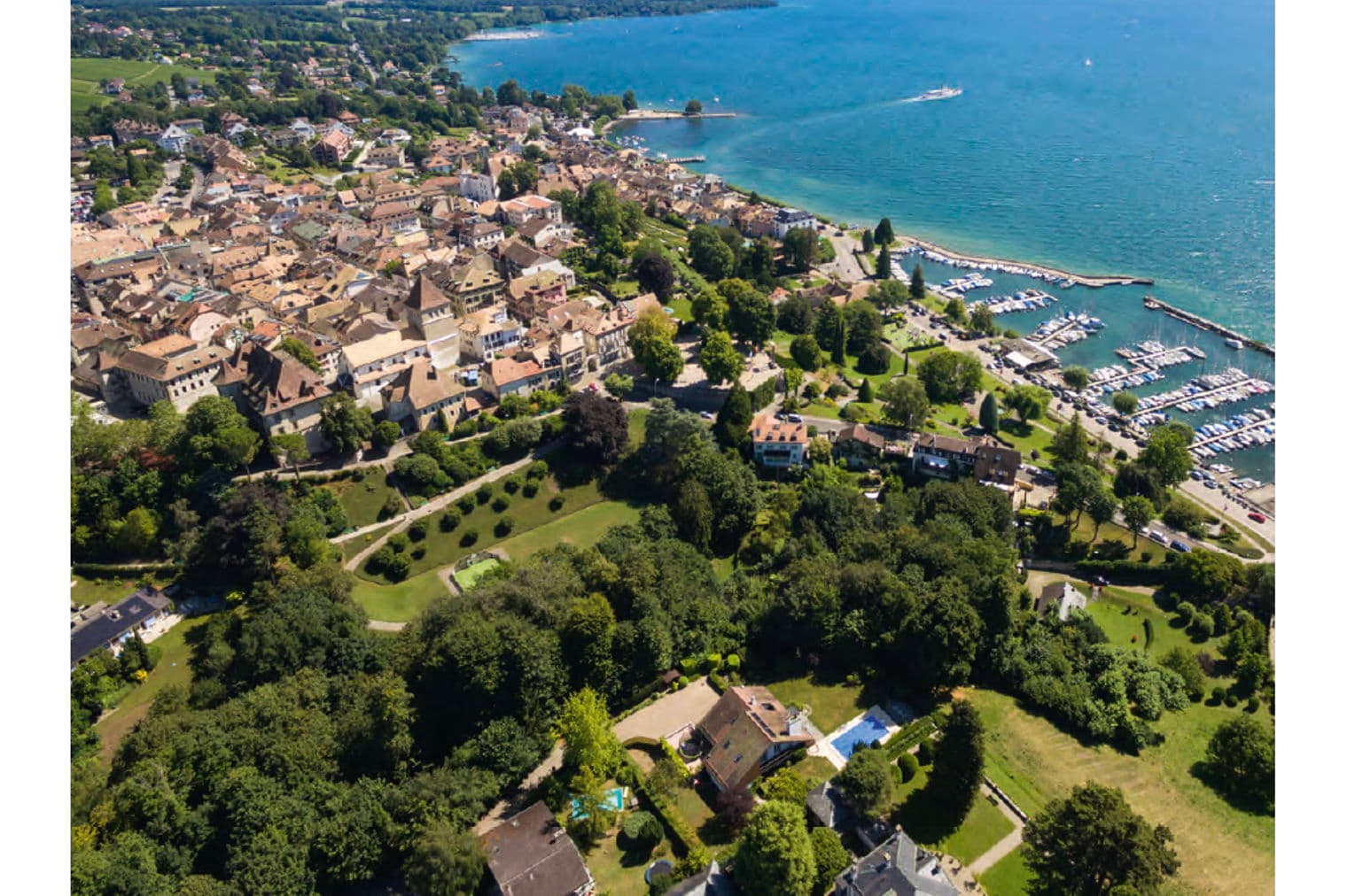 Engel & Völkers, vue aérienne de La Côte, Suisse. Lac bleu, bateaux et maisons aux toits rouges. Arbres verts et montagnes en arrière-plan.