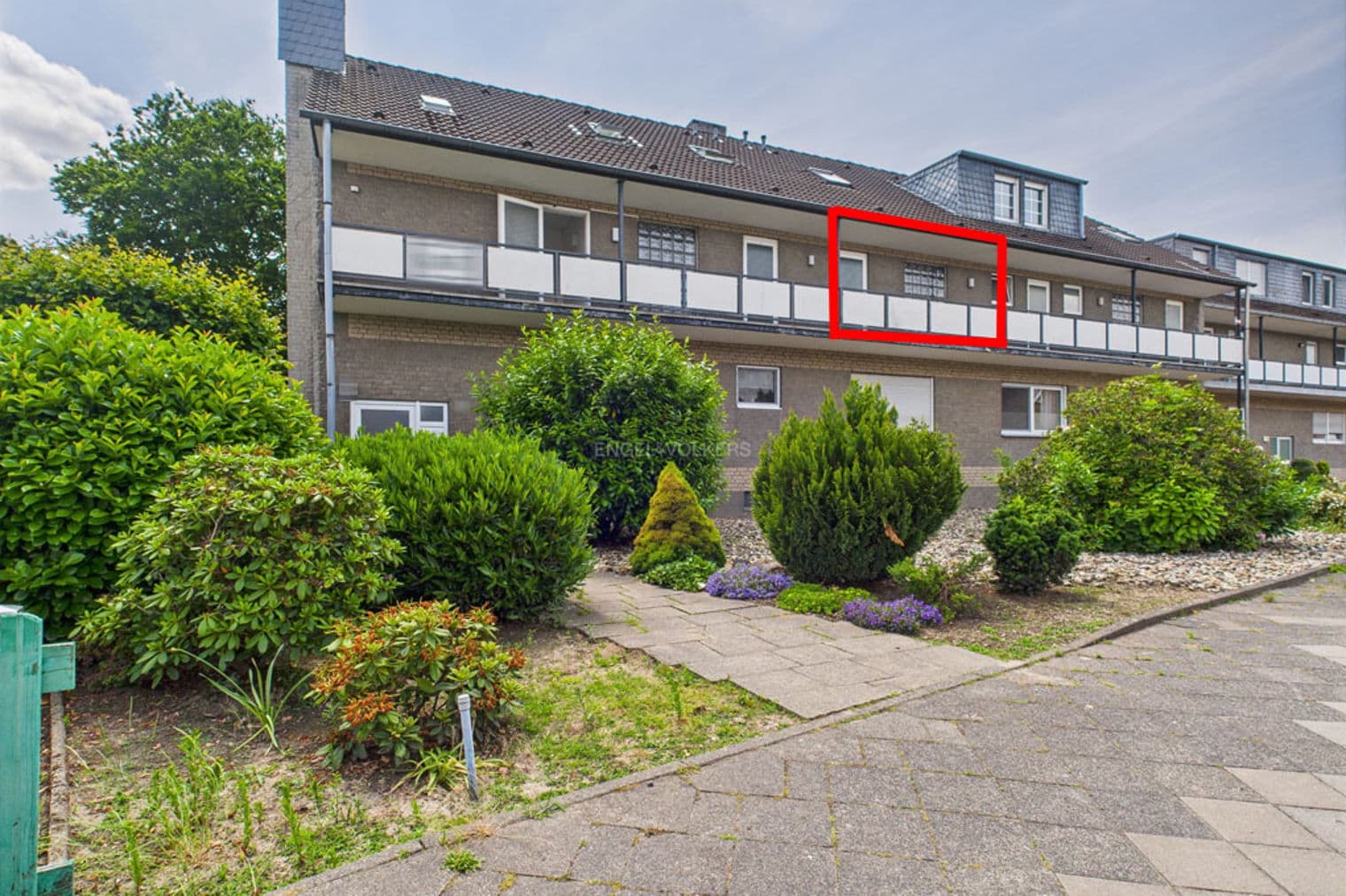 Three-story brick apartment building with white balconies and a red box highlighting one unit. Green bushes line the walkway.