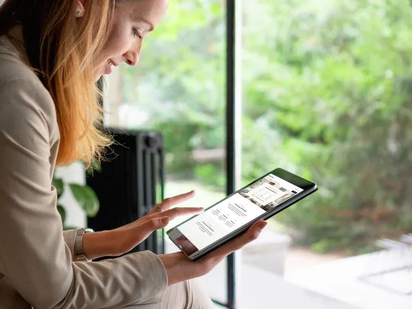 Person in a beige suit using a tablet near a window, displaying a webpage with text and images, with a blurred green background outside.
