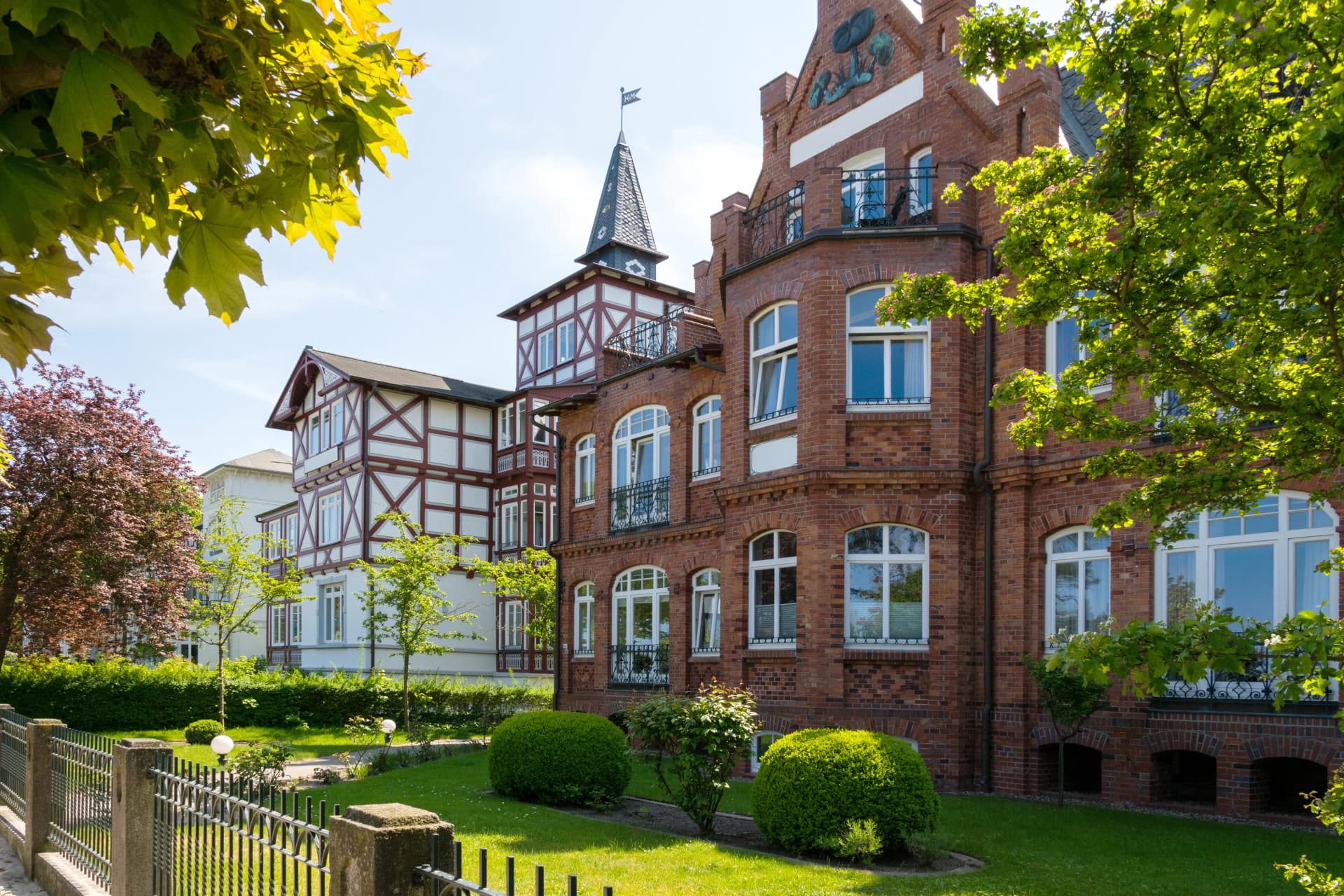 Red brick and white timbered buildings on a sunny day. Green trees frame the view of the architecture.