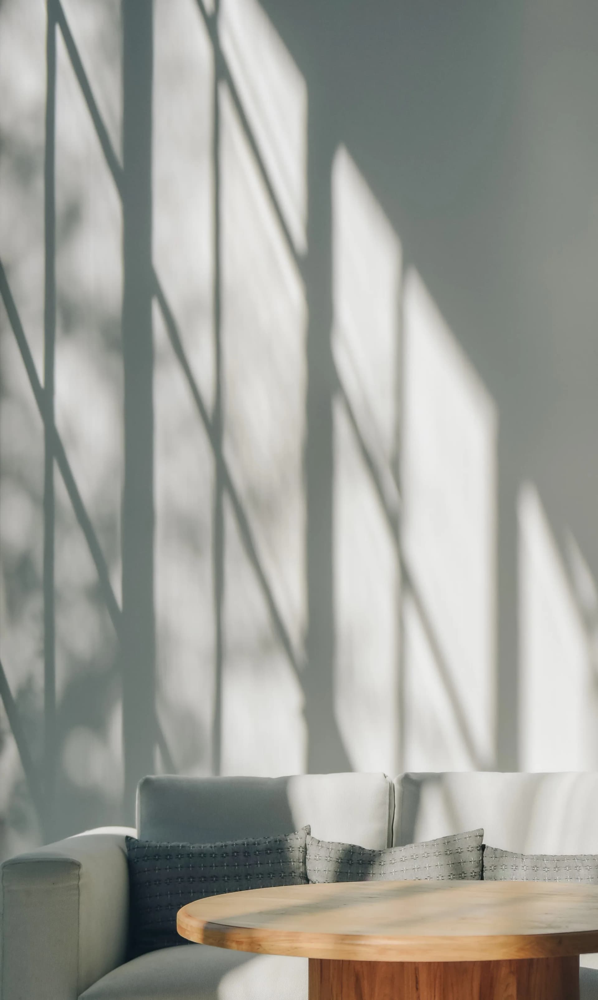 Sunlight streams through a window, casting shadows on a white wall above a gray sofa with pillows and a round wooden table.