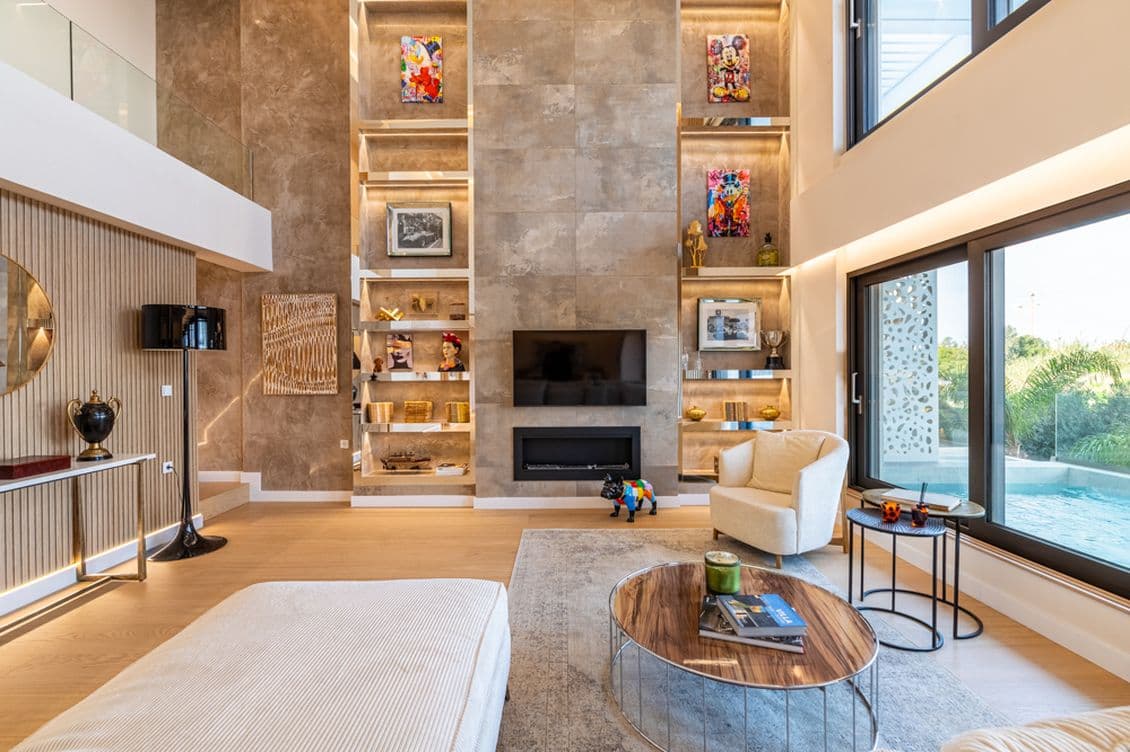 Ένα μοντέρνο σαλόνι με ψηλά ταβάνια, εντοιχισμένη μονάδα τοίχου με έργα τέχνης και διακόσμηση, μια τηλεόραση πάνω από το τζάκι, μεγάλα παράθυρα, ανοιχτόχρωμα έπιπλα, ένα στρογγυλό τραπέζι του καφέ και θέα σε μια εξωτερική πισίνα.