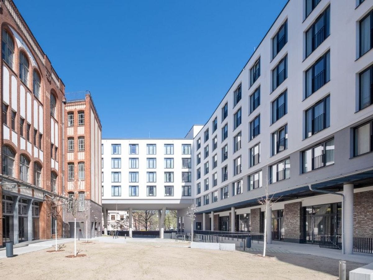 Modern and historic brick buildings form an open courtyard under a clear blue sky, with bare trees and green space in the center.