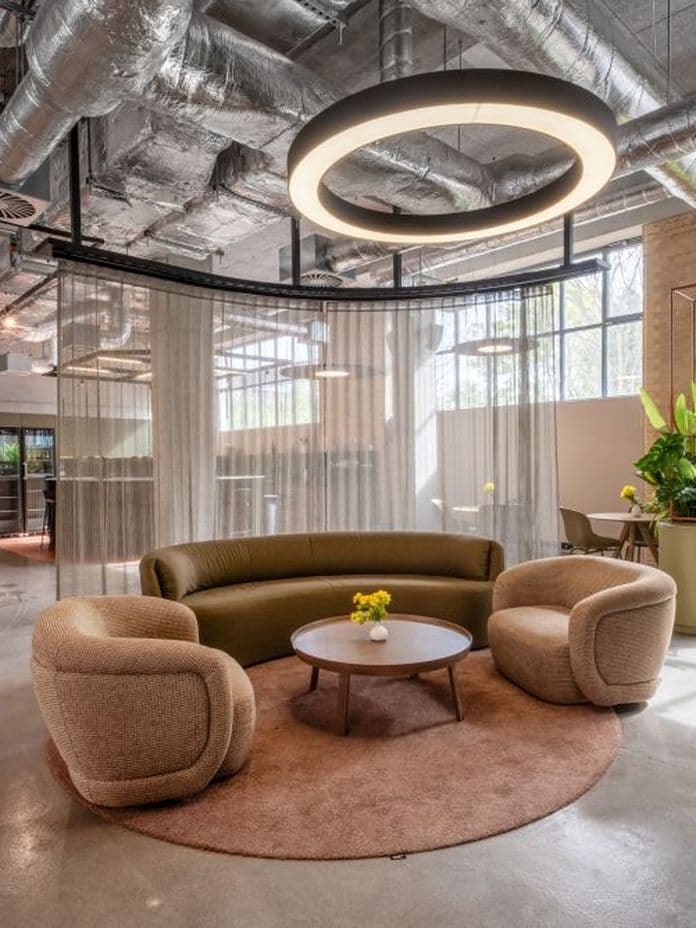 Modern office space with a round seating area, circular overhead light, large potted plant, exposed ductwork, and floor-to-ceiling windows.