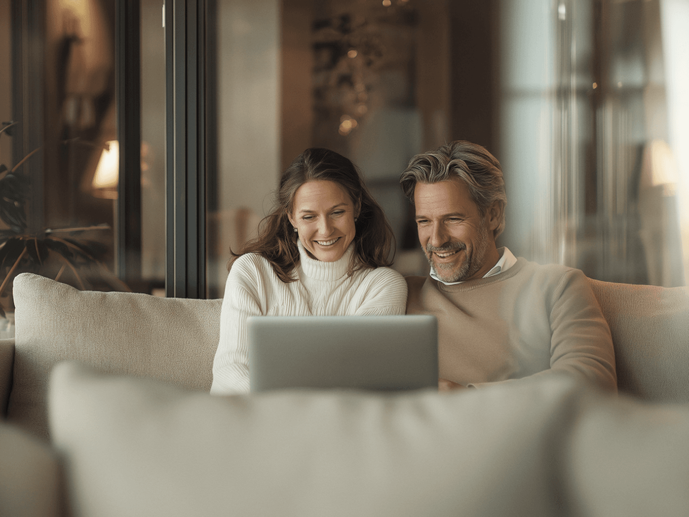 A smiling couple sitting on a sofa uses a laptop together, with a cozy home interior in the background.