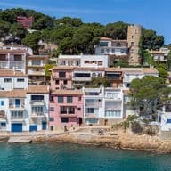 Vista costera de casas coloridas con techos de terracota en una costa rocosa, respaldada por una ladera verde y cubierta de árboles bajo un cielo azul.