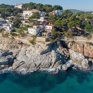 Coastal view of white villas with red tile roofs on a rocky cliff overlooking turquoise water. Green trees fill the background.