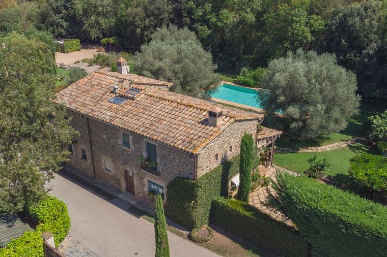 Aerial view of a stone house with a terracotta tile roof, surrounded by lush greenery and a blue swimming pool.