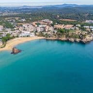 Vista aérea de una playa de arena y mar turquesa, con edificios y colinas verdes al fondo.