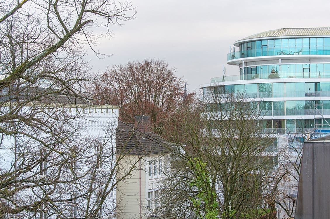 Modern glass building beside older white structures and bare trees under an overcast sky.