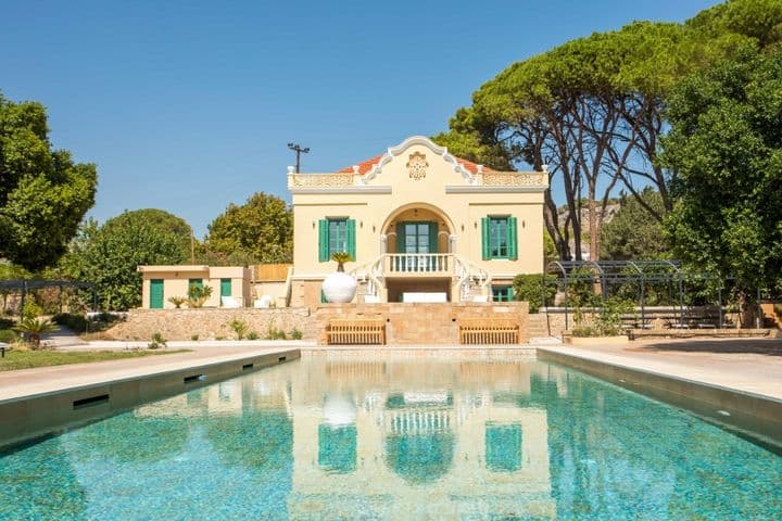 Elegant neoclassical villa with a private swimming pool, surrounded by lush greenery and tall pine trees under a clear blue sky.