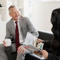 A man in a gray suit, holding a mug, smiles at a woman reading a magazine. They're seated in a modern, bright office space.