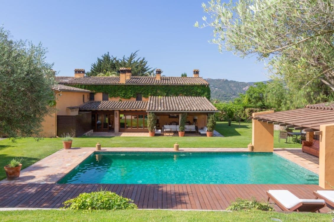 Außenansicht eines terrakottafarbenen Hauses mit Pool, grünem Rasen und Bäumen unter einem klaren blauen Himmel.