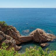 Costa rocosa con un puente de piedra sobre agua turquesa. Barcos salpican el mar azul bajo un cielo despejado. Una exuberante vegetación enmarca el primer plano.