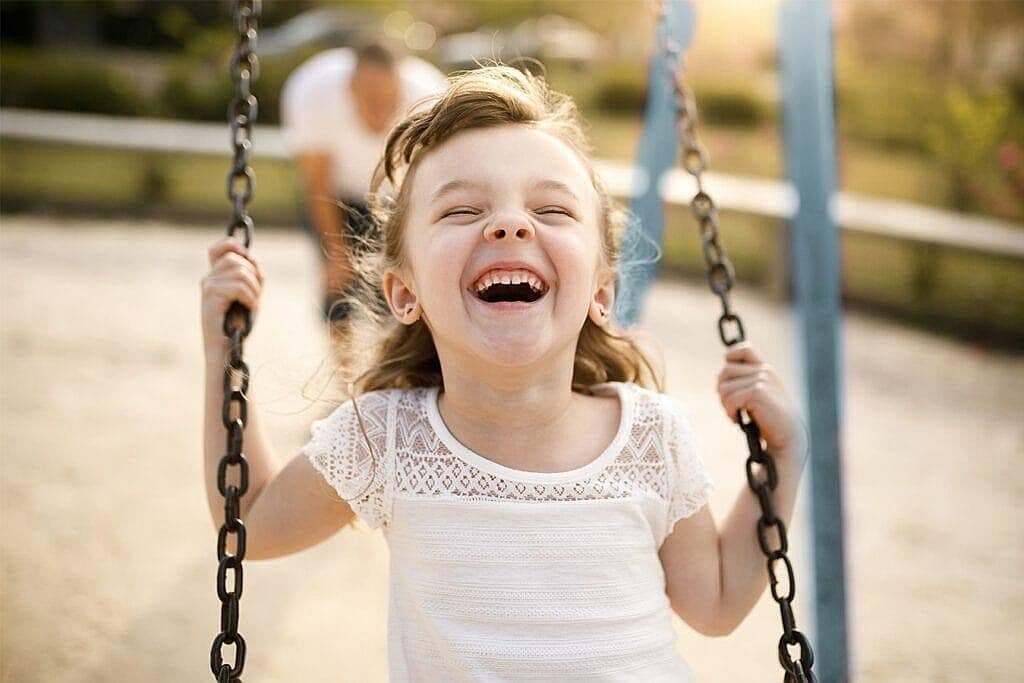 Una nena està molt contenta i somriu mentre es gronxa, es veu el seu pare darrere