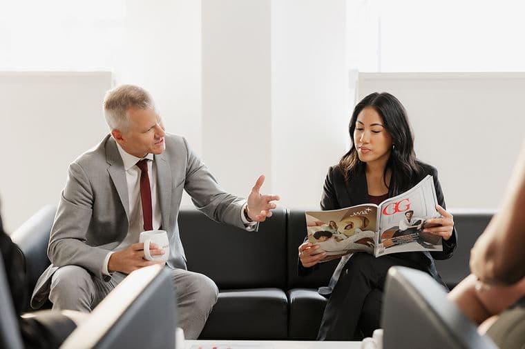 A man and woman in business attire sit on a couch. The man gestures while holding a mug, and the woman reads a magazine.
