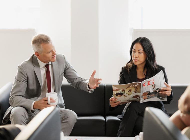 A man and woman in business attire sit on a couch. The man gestures while holding a mug, and the woman reads a magazine.