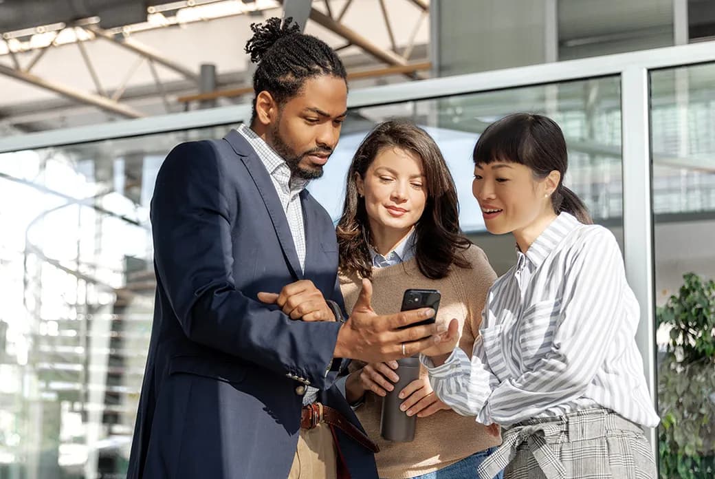 Three coworkers in a modern office review something on a smartphone together.