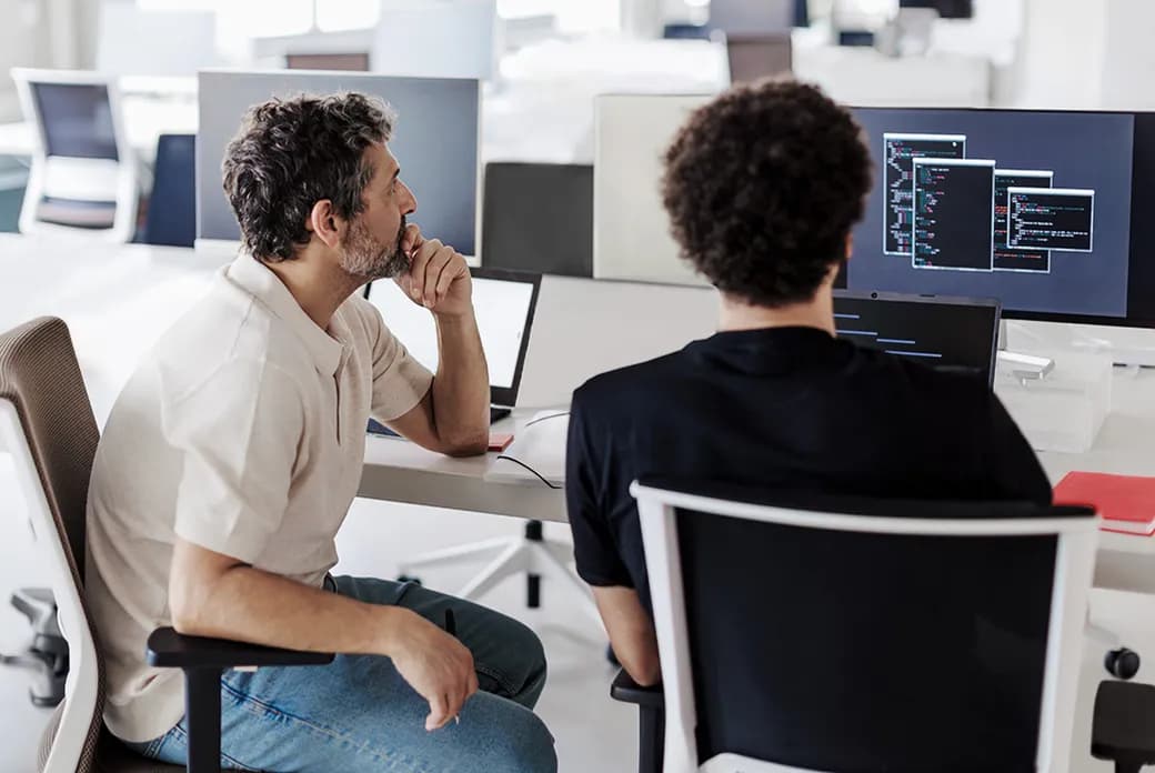 Two men collaborate at a bright, open-office desk with several monitors displaying code; one faces away while the other rests his chin in thought.