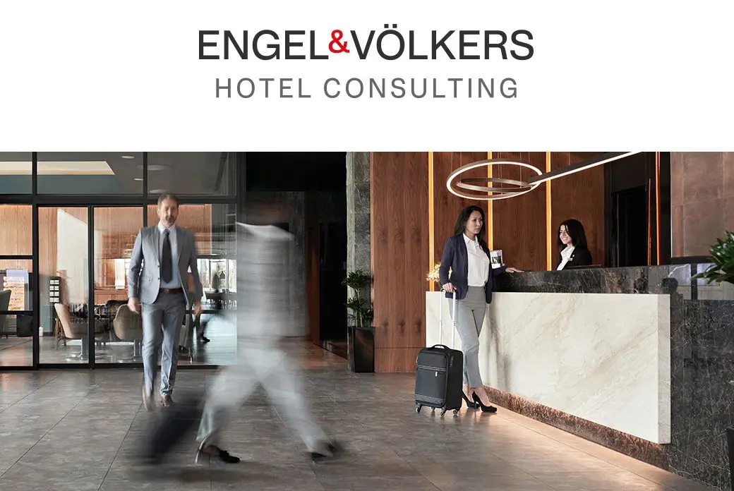 Hotel lobby with a woman checking in at the front desk, and a man walking through the lobby. Engel & Völkers logo at the top.