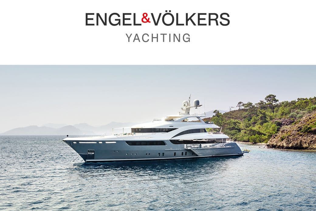 Engel & Völkers Yachting image: A large, gray and white yacht sits on calm, blue water near a green, tree-covered shoreline.