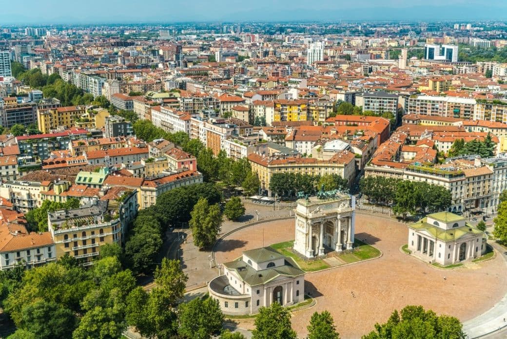 Aerial view of Milan featuring the Arco della Pace surrounded by historic buildings, tree-lined streets, and the city skyline in the background.