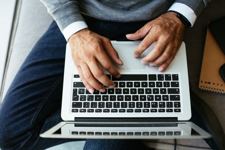 Uma pessoa digita em um laptop prateado enquanto está sentada. Ela veste jeans, um suéter cinza e um rastreador de atividades preto.