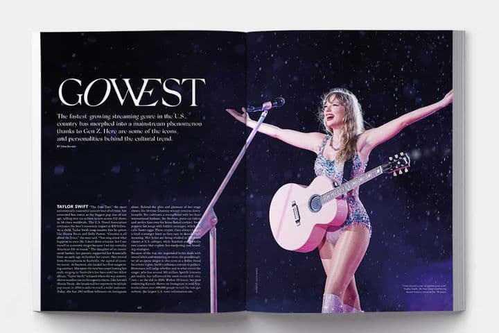 Taylor Swift on stage with a pink guitar, arms outstretched, wearing a sparkly blue outfit and silver boots.