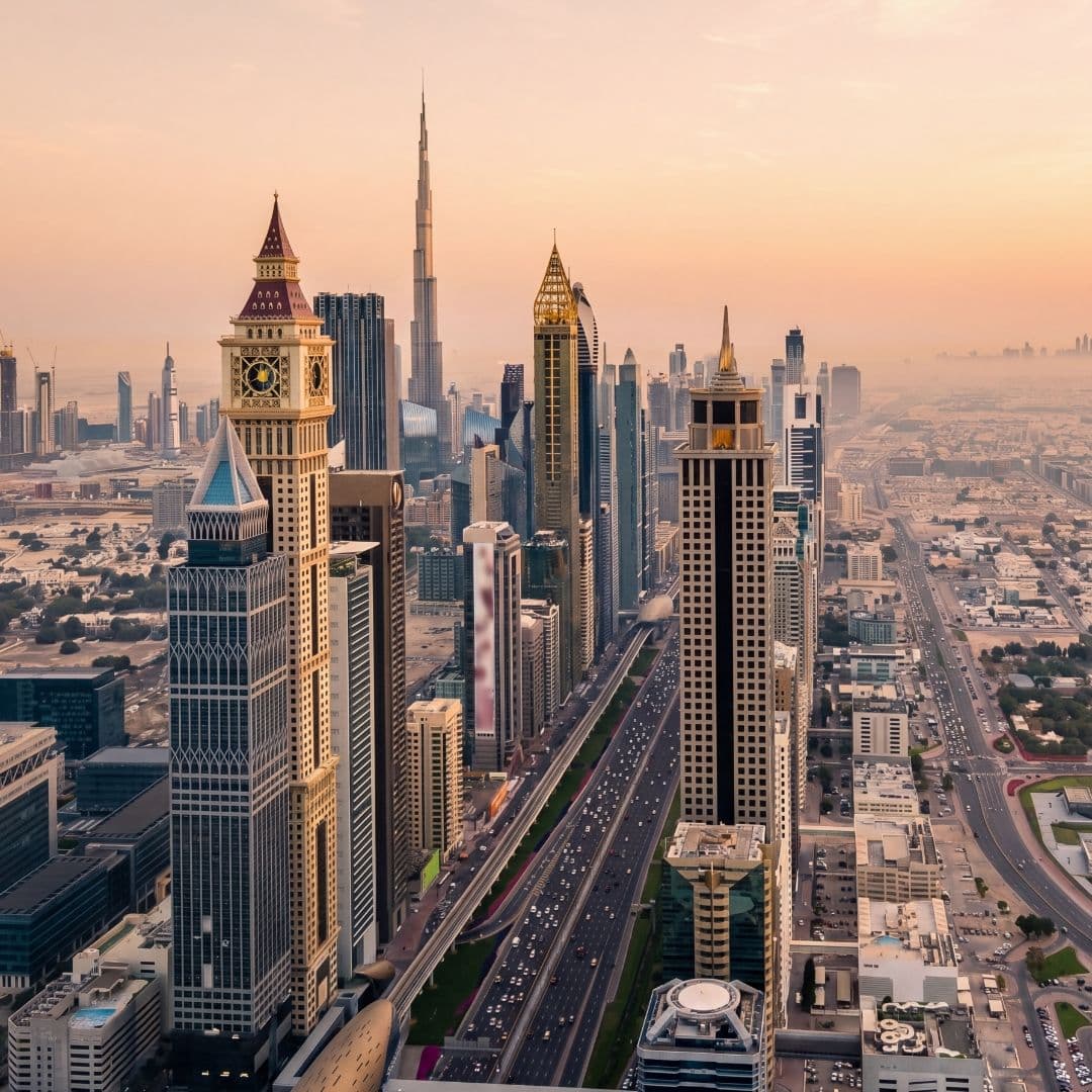 Aerial view of Dubai's skyline at sunset, featuring iconic skyscrapers and busy streets under a warm, hazy sky.