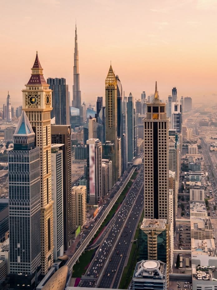 Aerial view of Dubai's skyline at sunset, featuring iconic skyscrapers and busy streets under a warm, hazy sky.