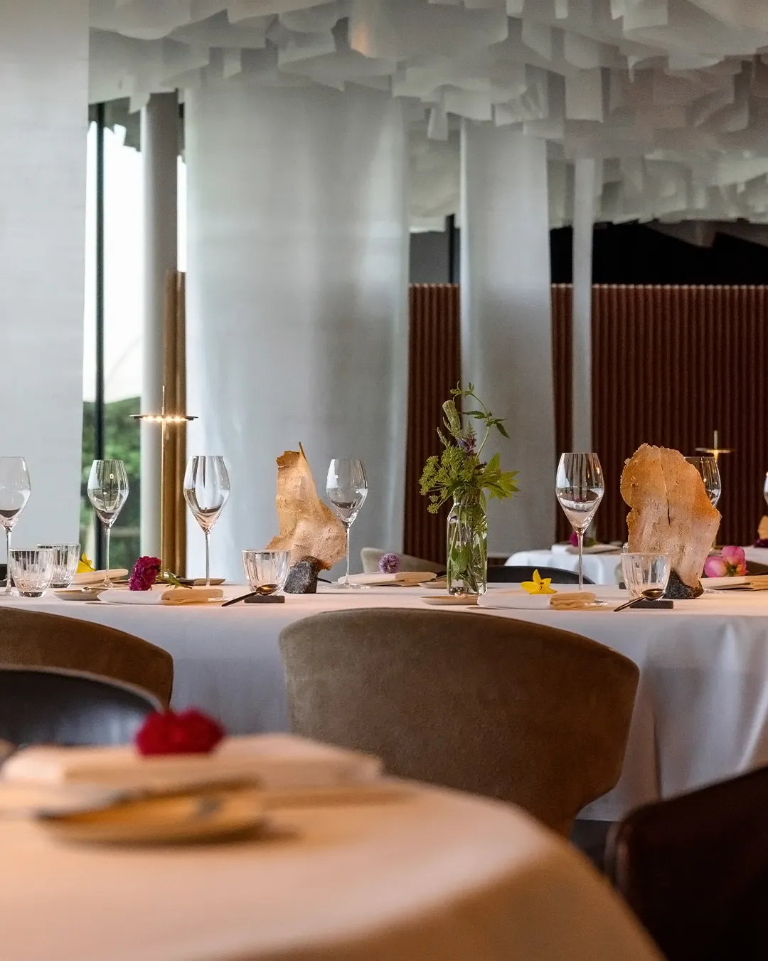 Elegant dining table set with wine glasses, plates, and a glass vase of greenery; rock-like centerpieces and a modern interior with white columns.