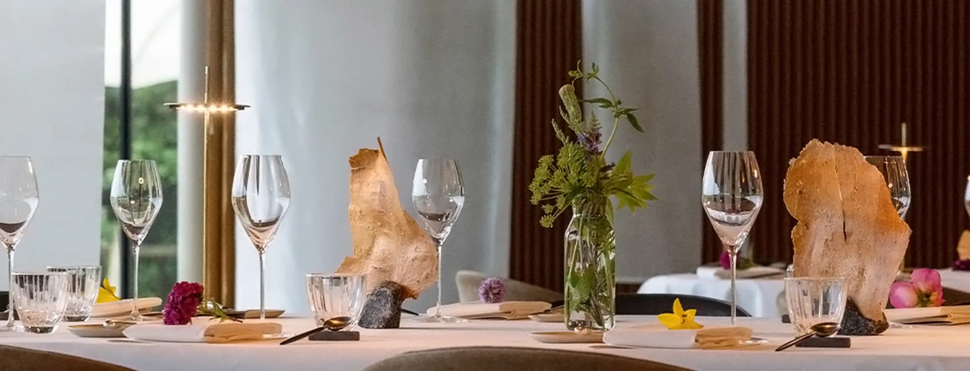 Elegant dining table set with wine glasses, plates, and a glass vase of greenery; rock-like centerpieces and a modern interior with white columns.