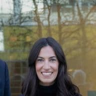 Drei Personen in Business-Kleidung stehen lächelnd vor einem Glasgebäude. Die Frau in der Mitte trägt einen hellblauen Anzug