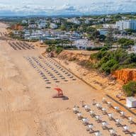 Aerial view of a sandy beach with umbrellas, a lifeguard post, and coastal cliffs. Nearby are residential buildings and lush greenery.