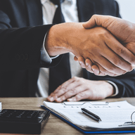 Zwei Personen schütteln sich über einem Vertrag auf einem Holzschreibtisch die Hände, auf dem sich ein Taschenrechner, ein Stift und Schlüssel befinden.