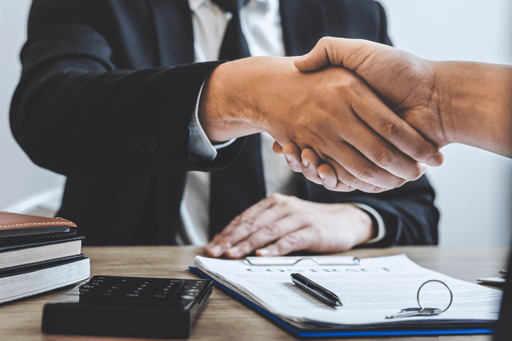 Zwei Personen schütteln sich über einem Vertrag auf einem Holzschreibtisch die Hände, auf dem sich ein Taschenrechner, ein Stift und Schlüssel befinden.