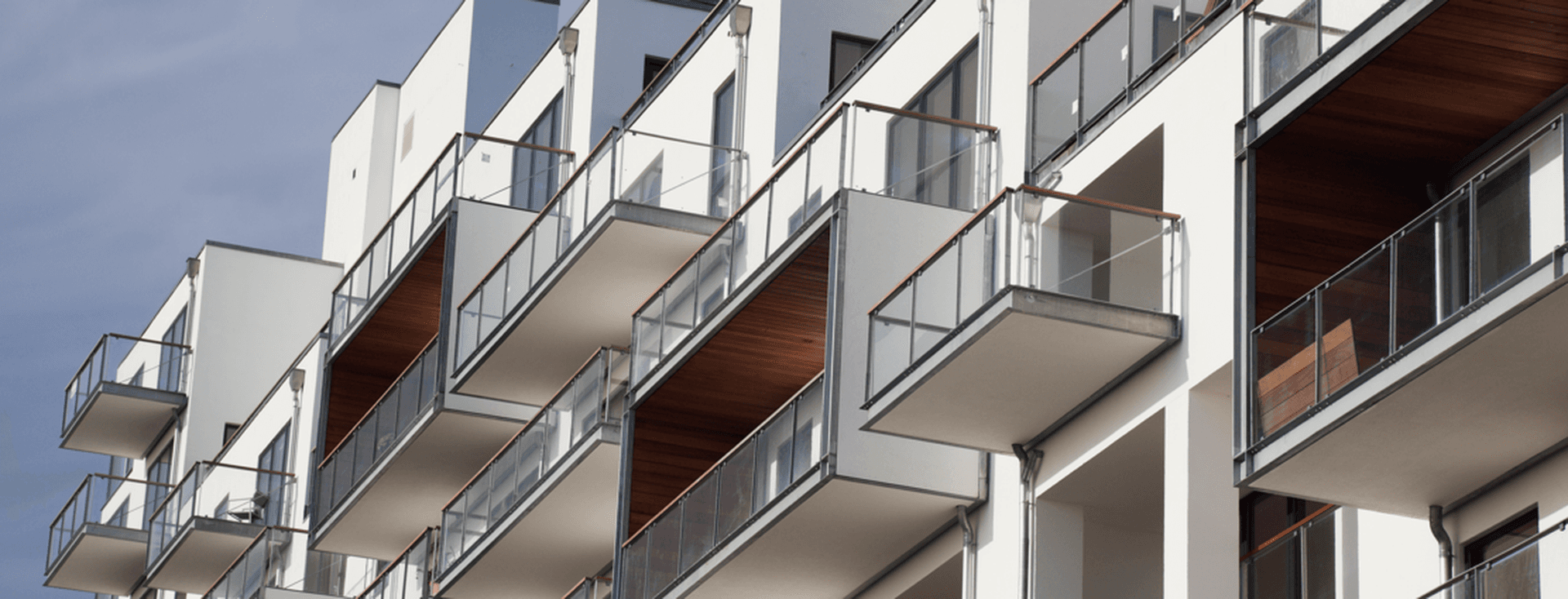Edifici d'apartaments modern blanc amb balcons de vidre i sostres de fusta amb un cel blau de fons.