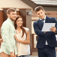 Ein Immobilienmakler in einem blauen Anzug zeigt einem lächelnden Paar vor einem Haus ein Tablet.