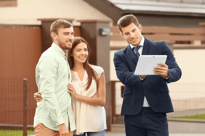 Un agent immobiliari amb un vestit blau mostra una tauleta a una parella somrient davant d'una casa.